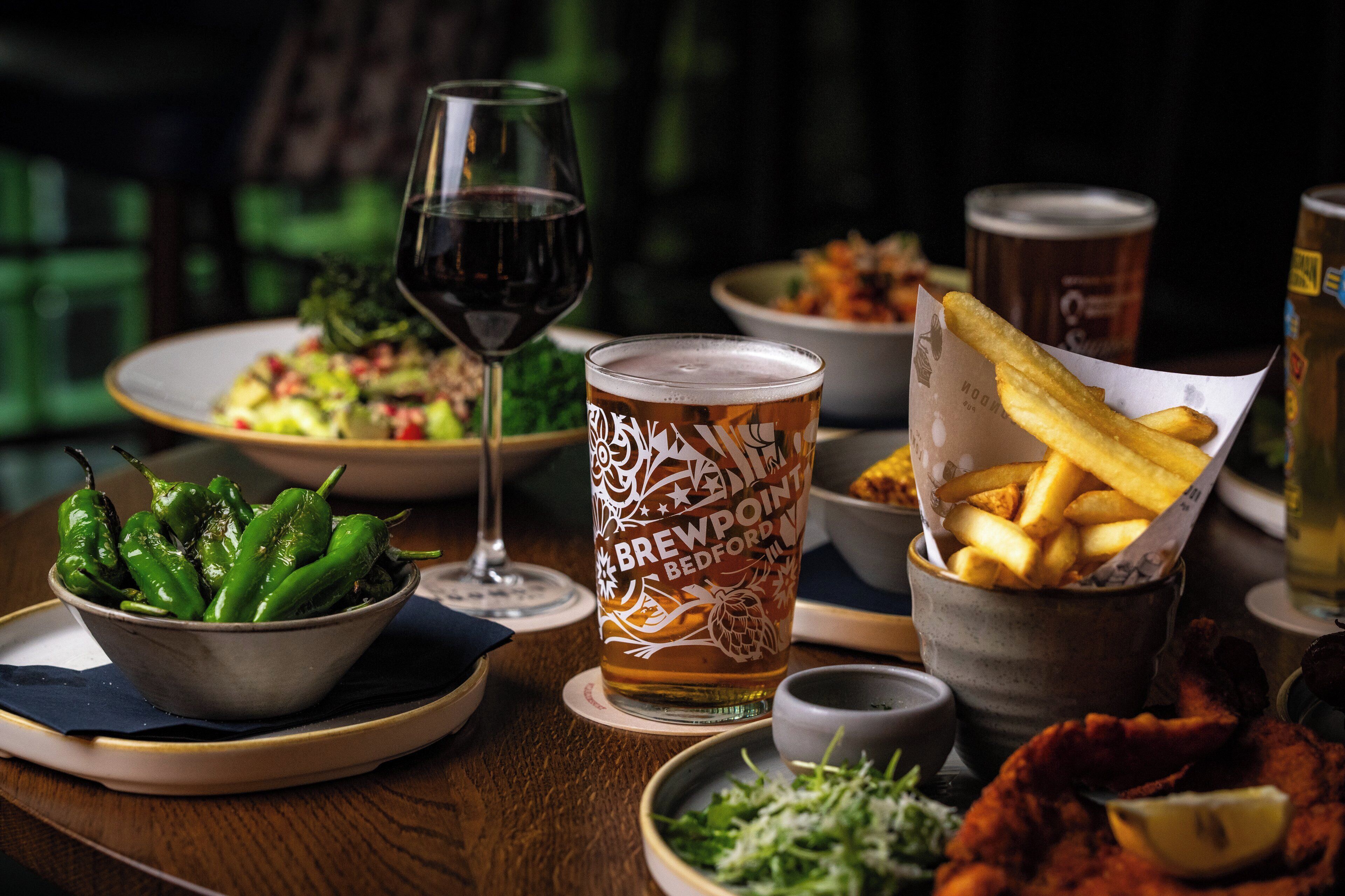 A table with a bowl of green peppers, a glass of beer, a plate of food, and a basket of fries.