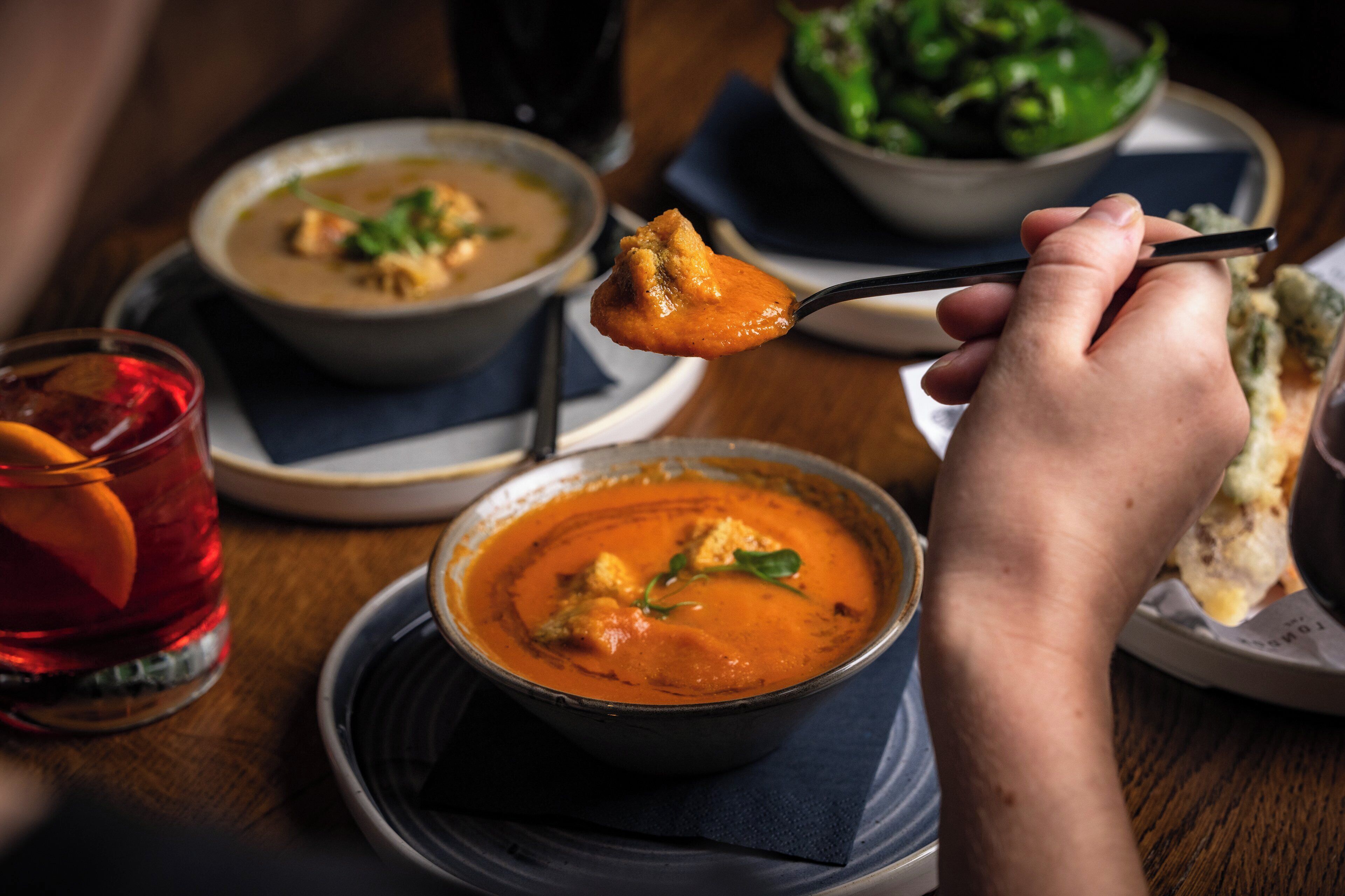 Eine Person hält einen Löffel über einer Schüssel Suppe, ein Glas rotes Getränk und einen Teller mit Essen stehen auf einem Tisch.