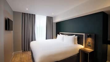 A modern hotel room with a large bed, a flat-screen TV mounted on the wall, and a bedside table with a lamp.