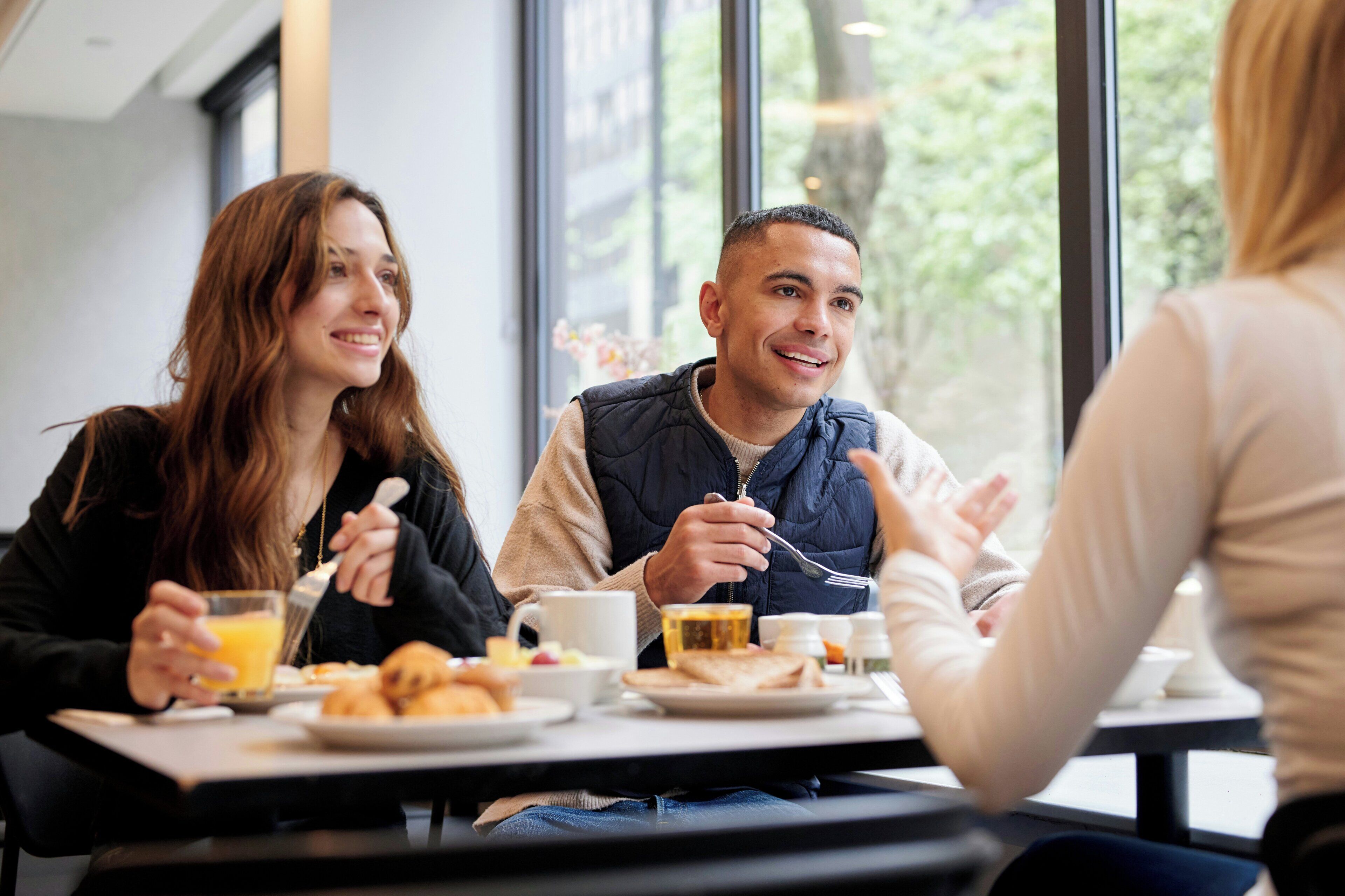 Tres personas comiendo en una mesa con alimentos para el desayuno, vistas al exterior y un interior moderno.