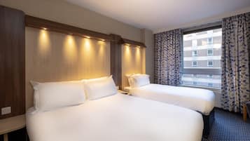 Habitación de hotel con dos camas, cabecero de madera, una ventana con cortinas estampadas y vista a un edificio.
