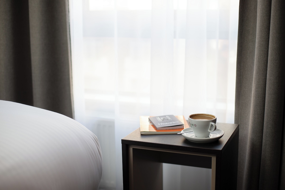 A neatly made bed with a white comforter, a small bedside table with a cup of coffee, a notebook, and a smartphone.