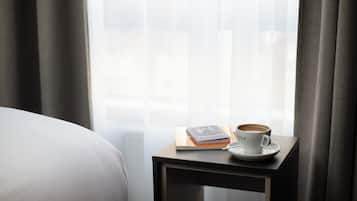 Una cama bien hecha con una colcha blanca, una mesita de noche con una taza de café, un cuaderno y un teléfono inteligente.