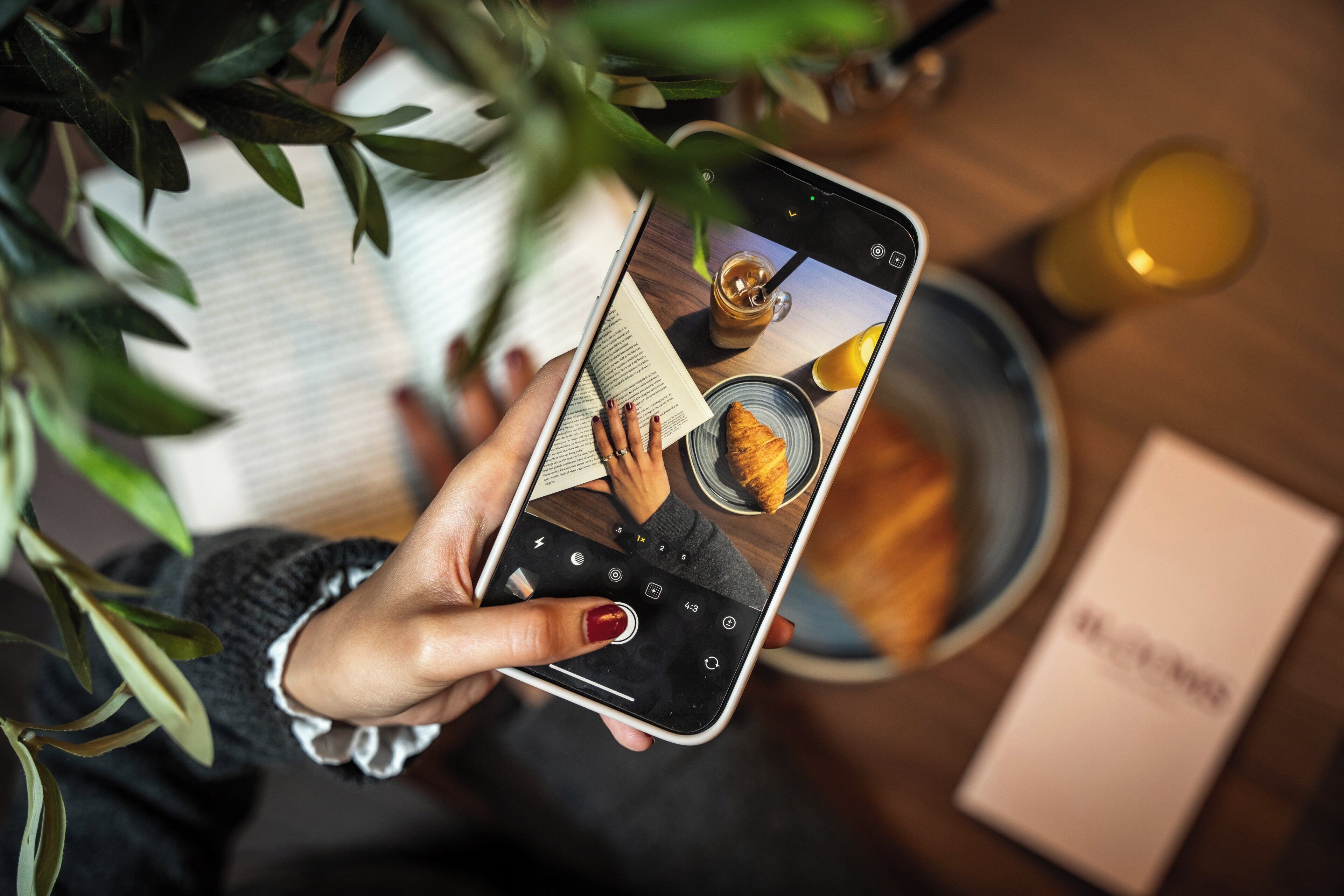 A person holding a smartphone with a camera interface, a book, and a plate with a croissant.