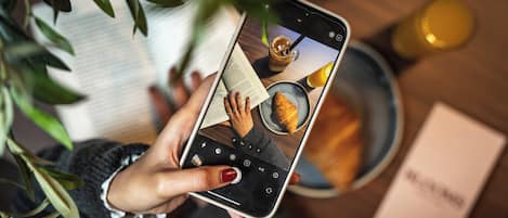 A person holding a smartphone with a camera interface, a book, and a plate with a croissant.
