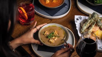 Una persona sosteniendo un tazón de sopa con una cuchara, un vaso de bebida roja, y un plato con pan y una taza de líquido negro.