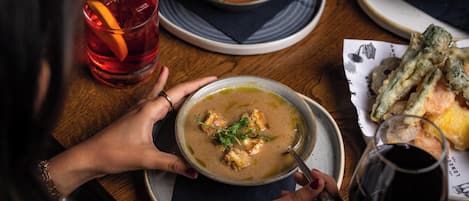 A person holding a bowl of soup with a spoon, a glass of red drink, and a plate with bread and a cup of black liquid.