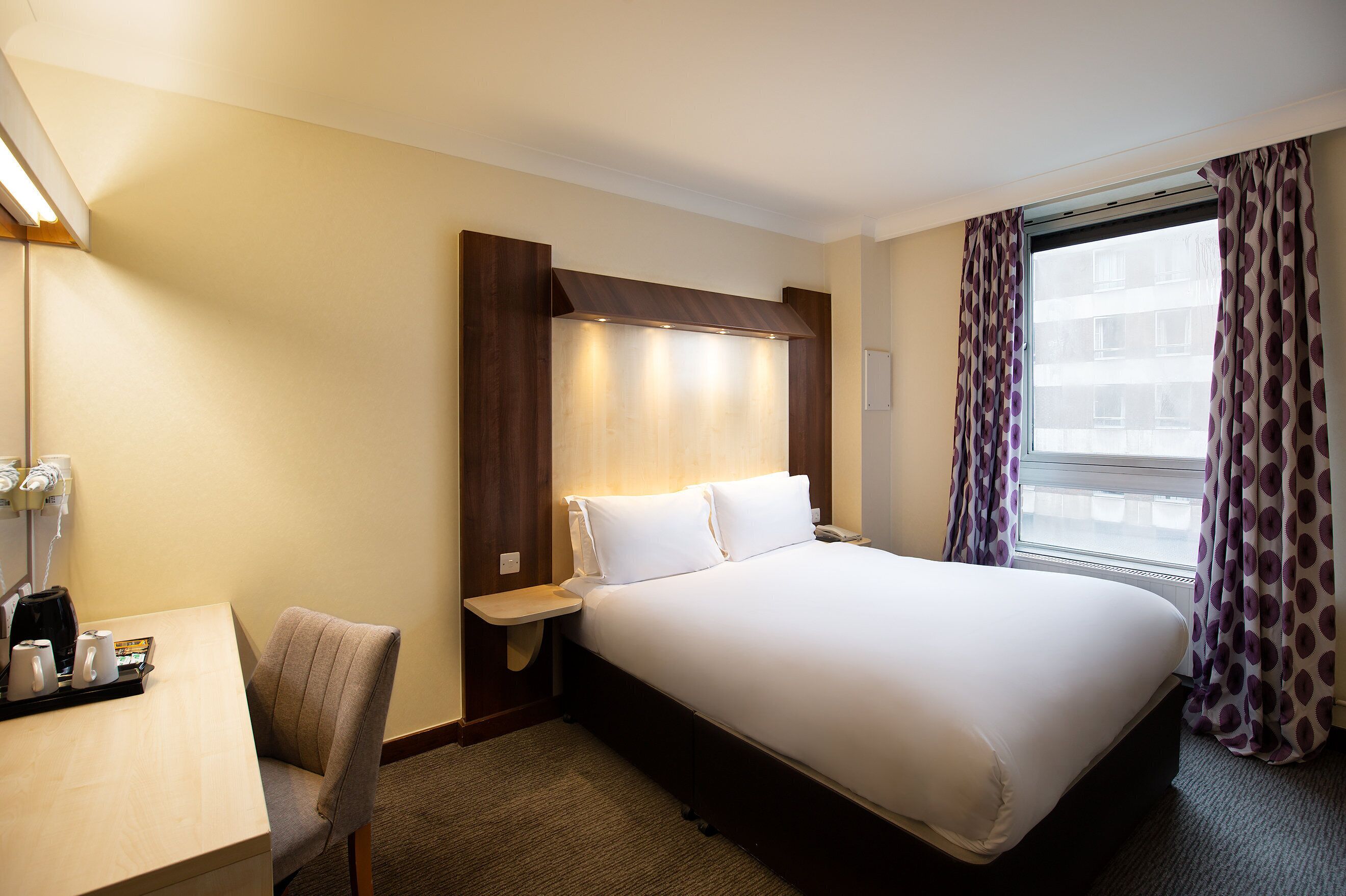 A hotel room with a bed, a desk with a chair, a bedside table, and a window with curtains.