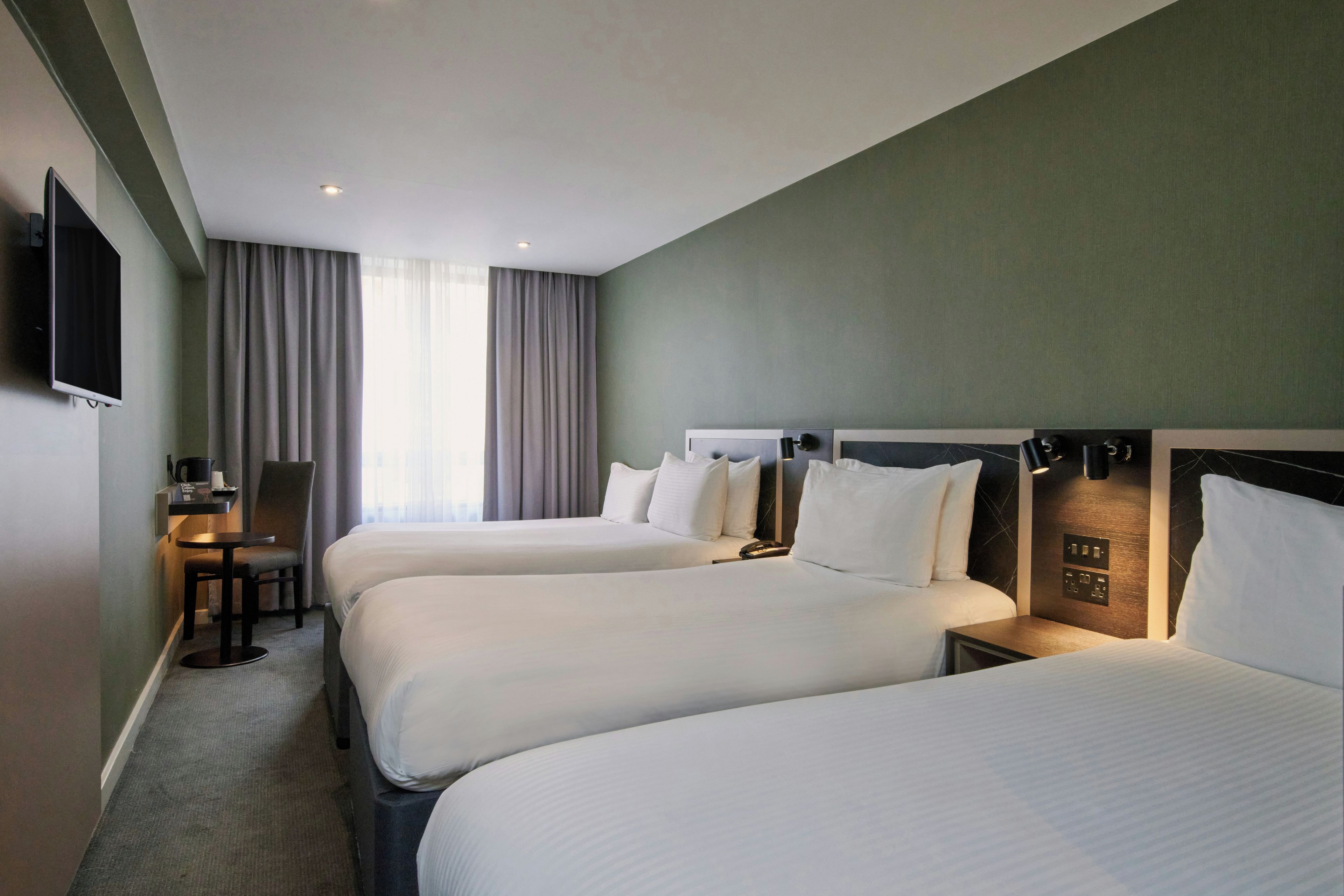 Ein Hotelzimmer mit zwei Betten, einem Schreibtisch, einem Stuhl, einem Fernseher und einem Fenster mit Vorhängen.