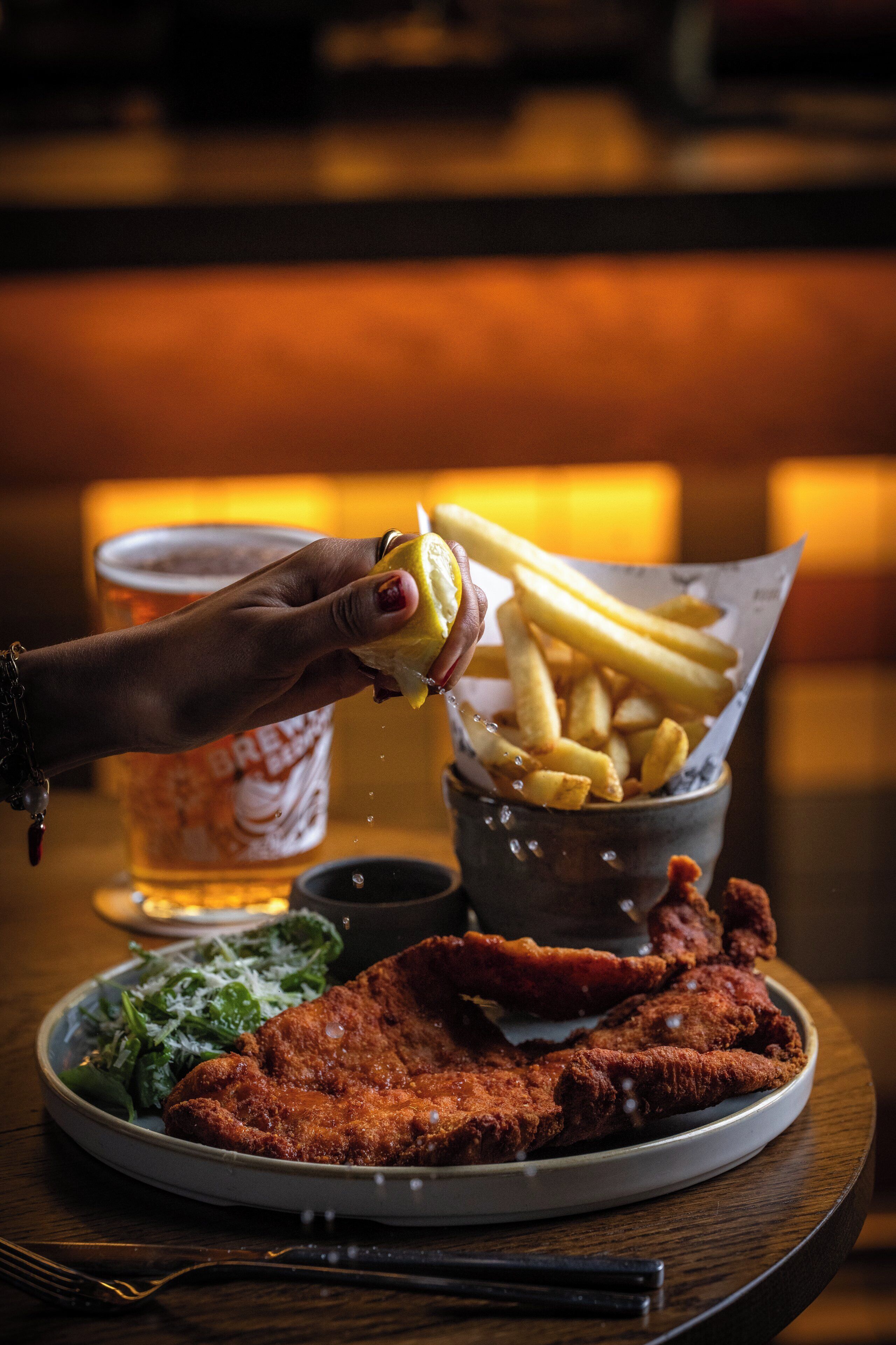 A plate with fried chicken, fries, and salad, a glass of beer, and a hand squeezing a lemon over the fries.