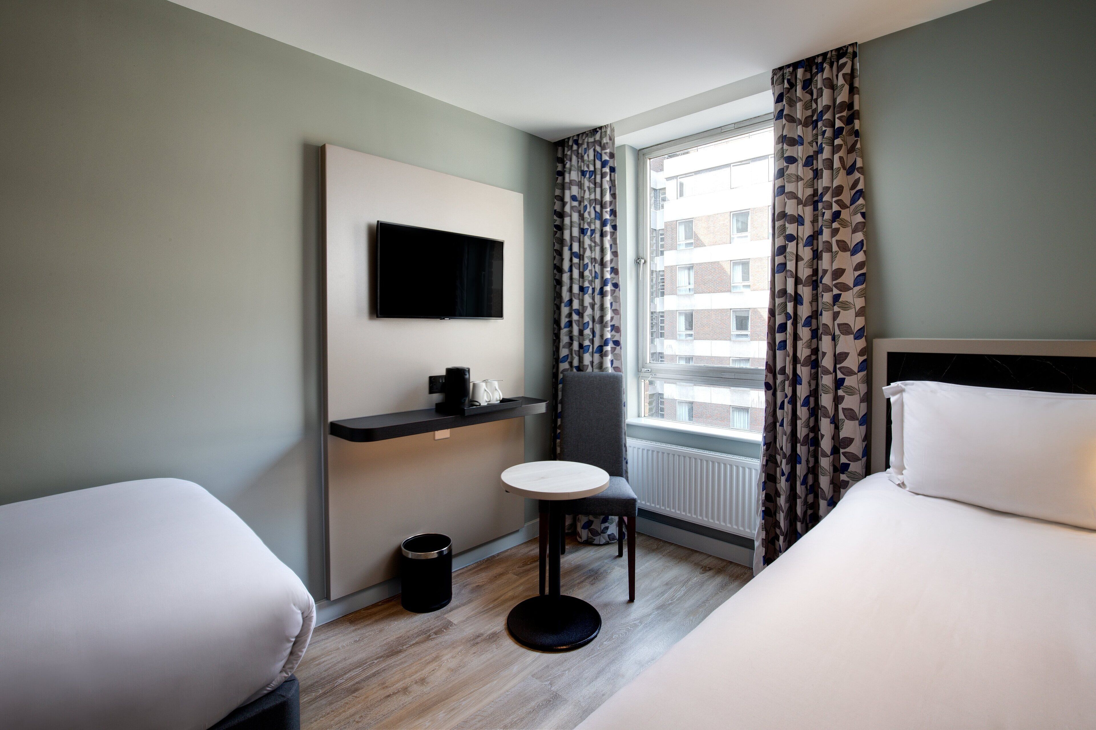 Une chambre d’hôtel avec deux lits, une petite table, un téléviseur, une fenêtre avec des rideaux et un radiateur.