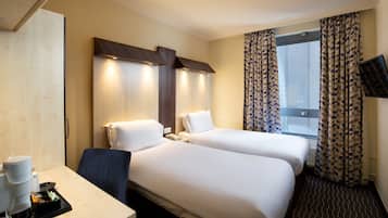 Ein Hotelzimmer mit zwei Betten, einem Schreibtisch mit Kaffeemaschine, einem Fernseher und einem Fenster mit gemusterten Vorhängen.