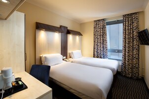 A hotel room with two beds, a desk with a coffee maker, a TV, and a window with patterned curtains.