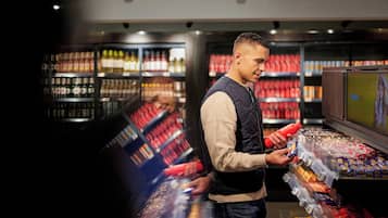 A person in a vest and sweater holding a red bottle, standing in a store aisle with shelves stocked with various products.