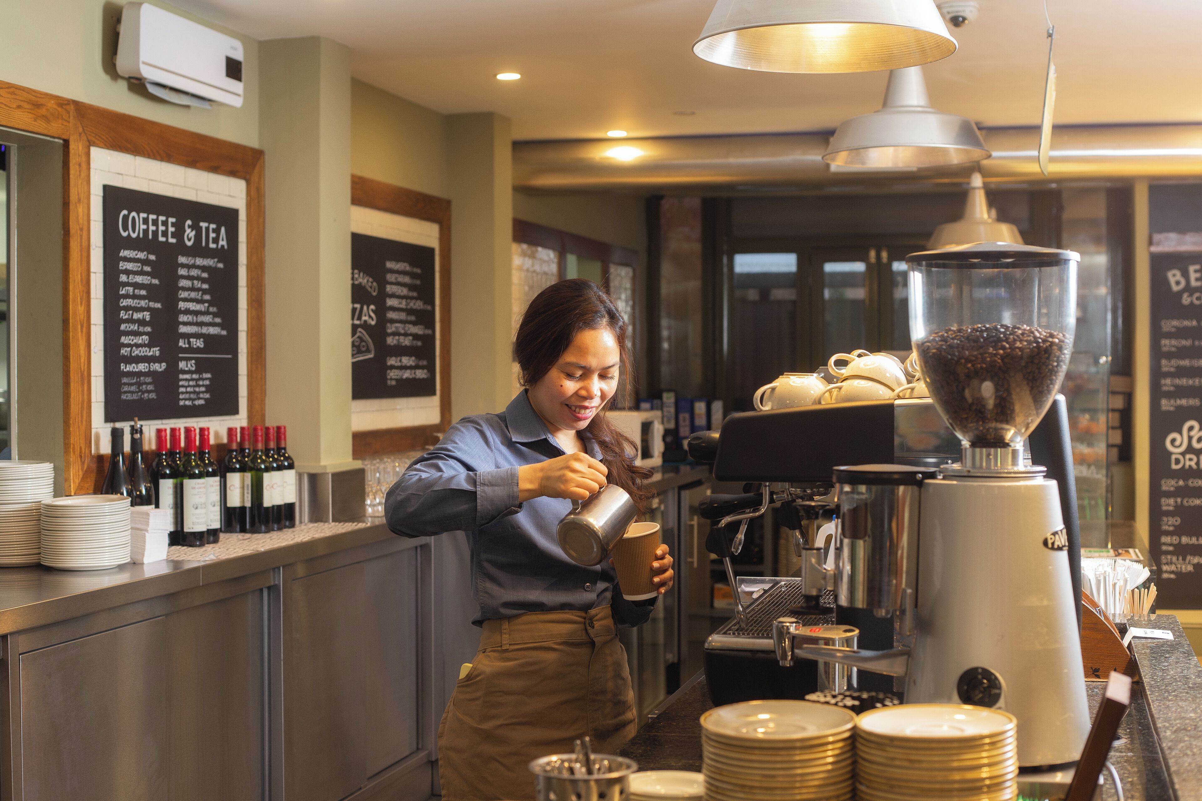 Un barista preparando una bebida en la barra de una cafetería, con una máquina de café, carteles de menú y diversos artículos relacionados con el café.