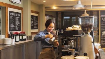 A barista preparing a drink at a coffee shop counter with a coffee machine, menu boards, and various coffee-related items.