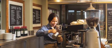 Un barista preparando una bebida en la barra de una cafetería, con una máquina de café, carteles de menú y diversos artículos relacionados con el café.