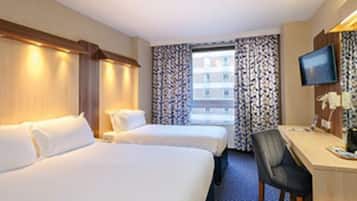 A hotel room with two beds, a desk with a chair, a television, and a window with patterned curtains.