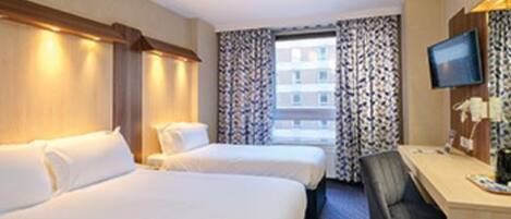 A hotel room with two beds, a desk with a chair, a television, and a window with patterned curtains.