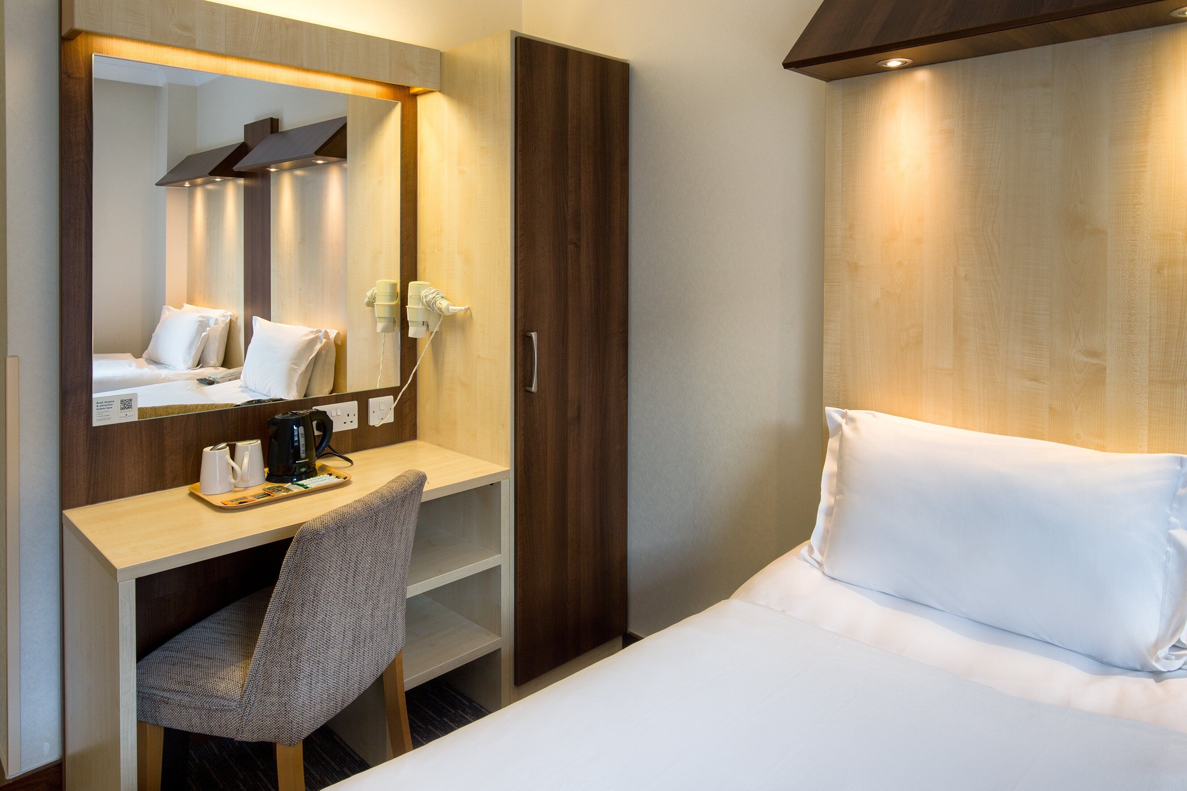A hotel room with a bed, a desk with a chair, a mirror, and a wardrobe.