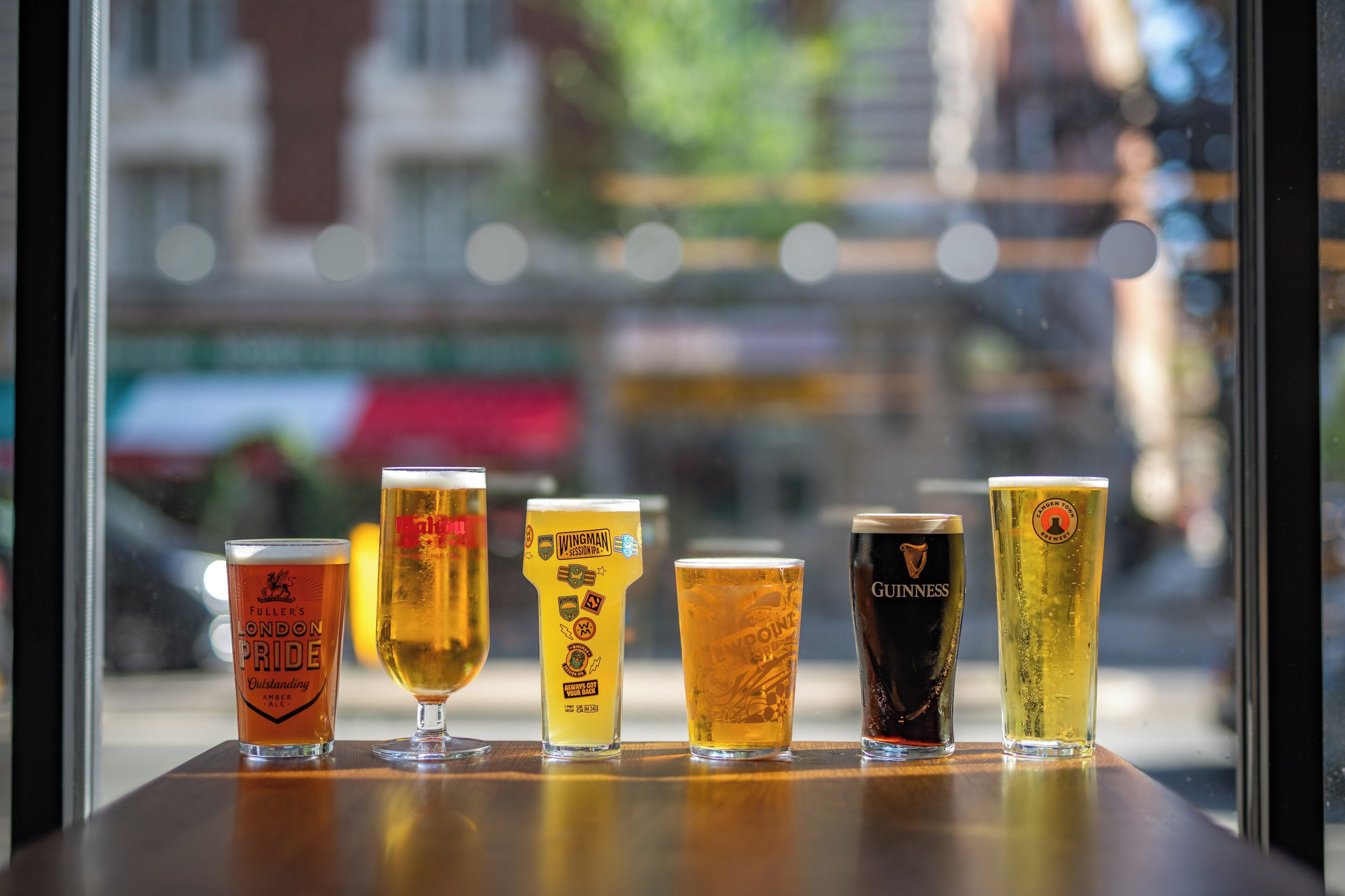 Une rangée de six verres à bière est posée sur une table, chacun portant une étiquette différente, devant une fenêtre donnant sur un paysage urbain flou.