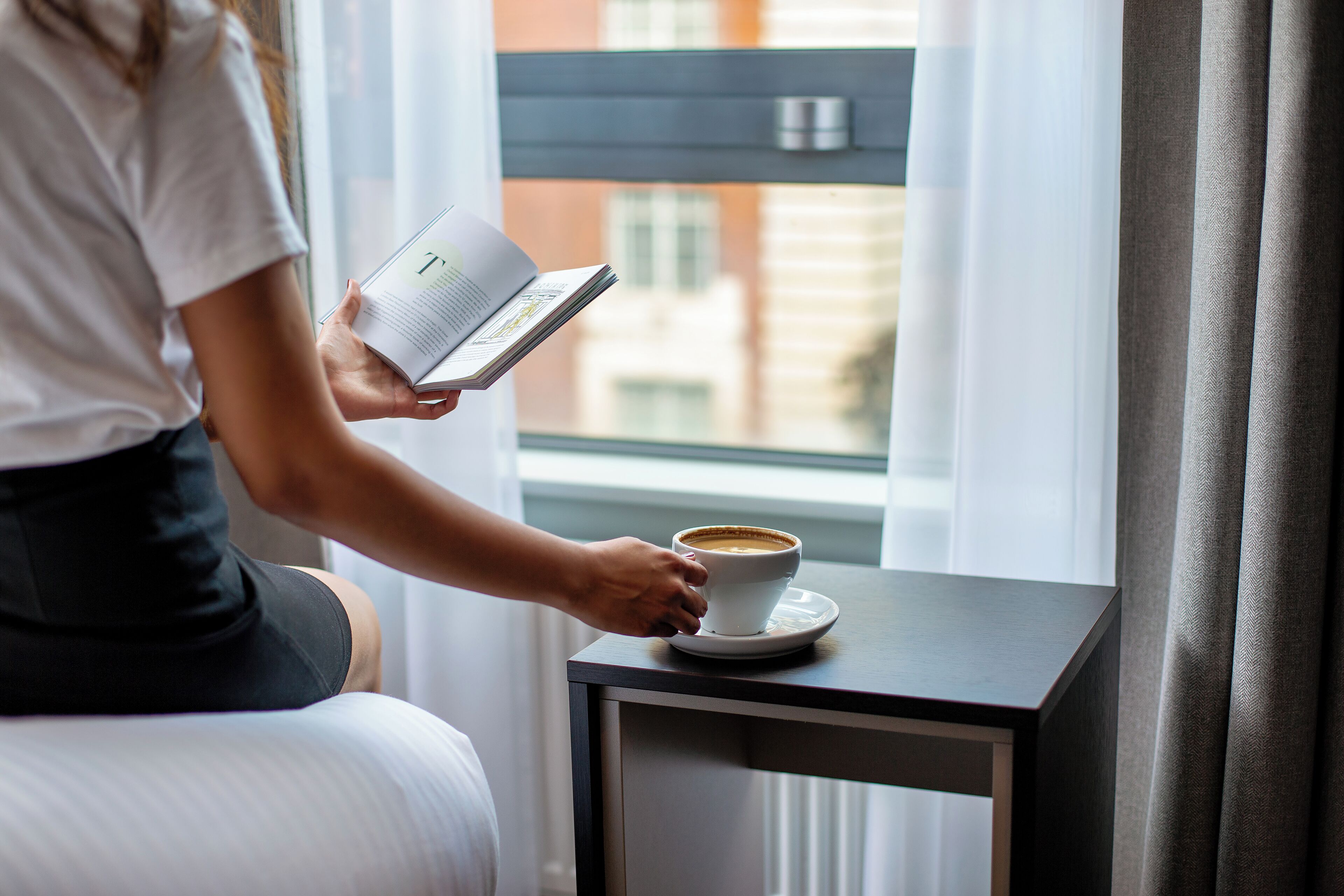 Una persona leyendo un libro junto a una taza de café sobre una pequeña mesa cerca de una ventana.