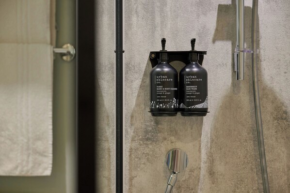 A bathroom with two black bottles labeled 'urban skincare co.' on a wall-mounted holder, a showerhead, and a towel rack. - Royal National Hotel (London)