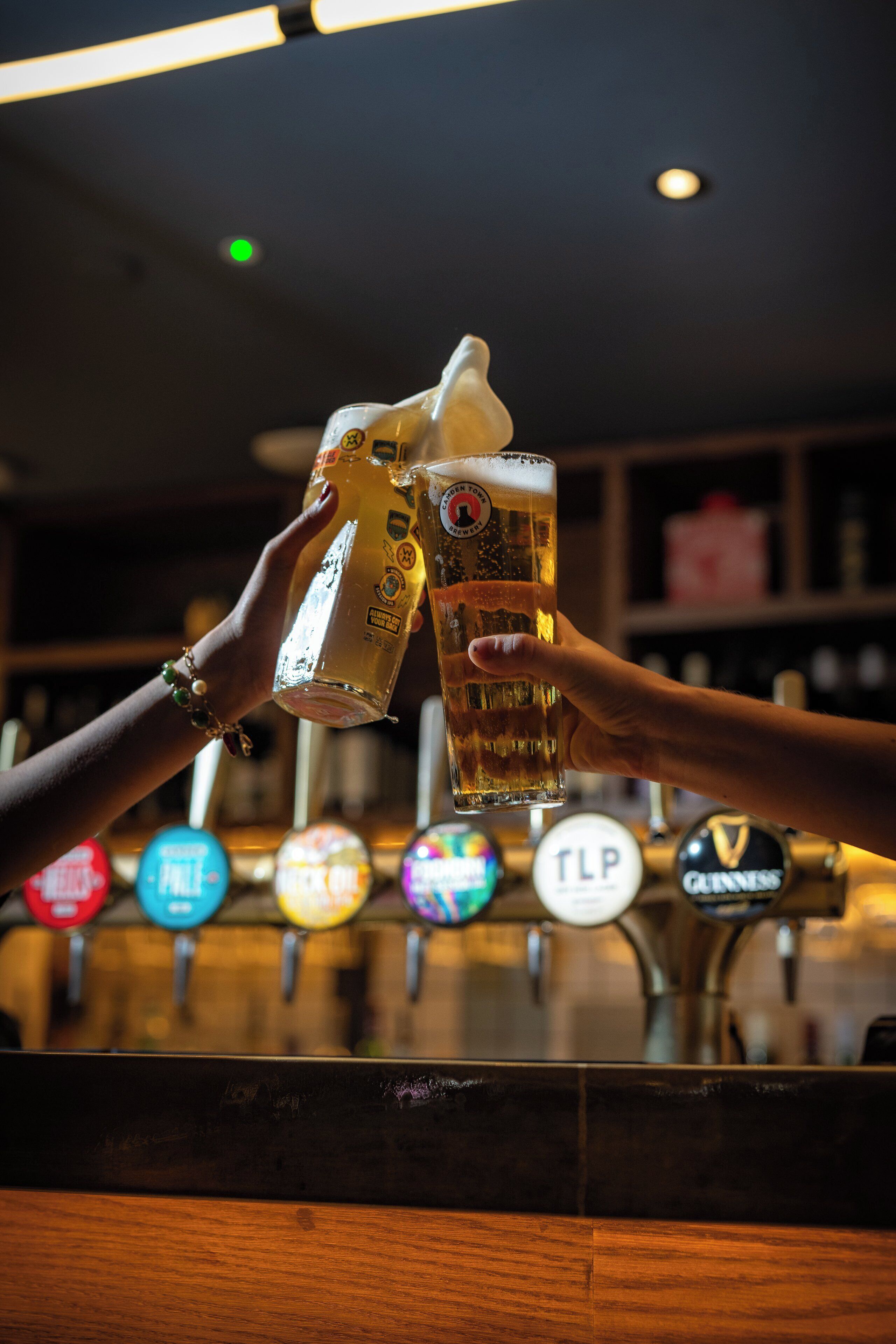 A bar with two people toasting with beer glasses. Beer taps with logos like TPL and Guinness are visible.