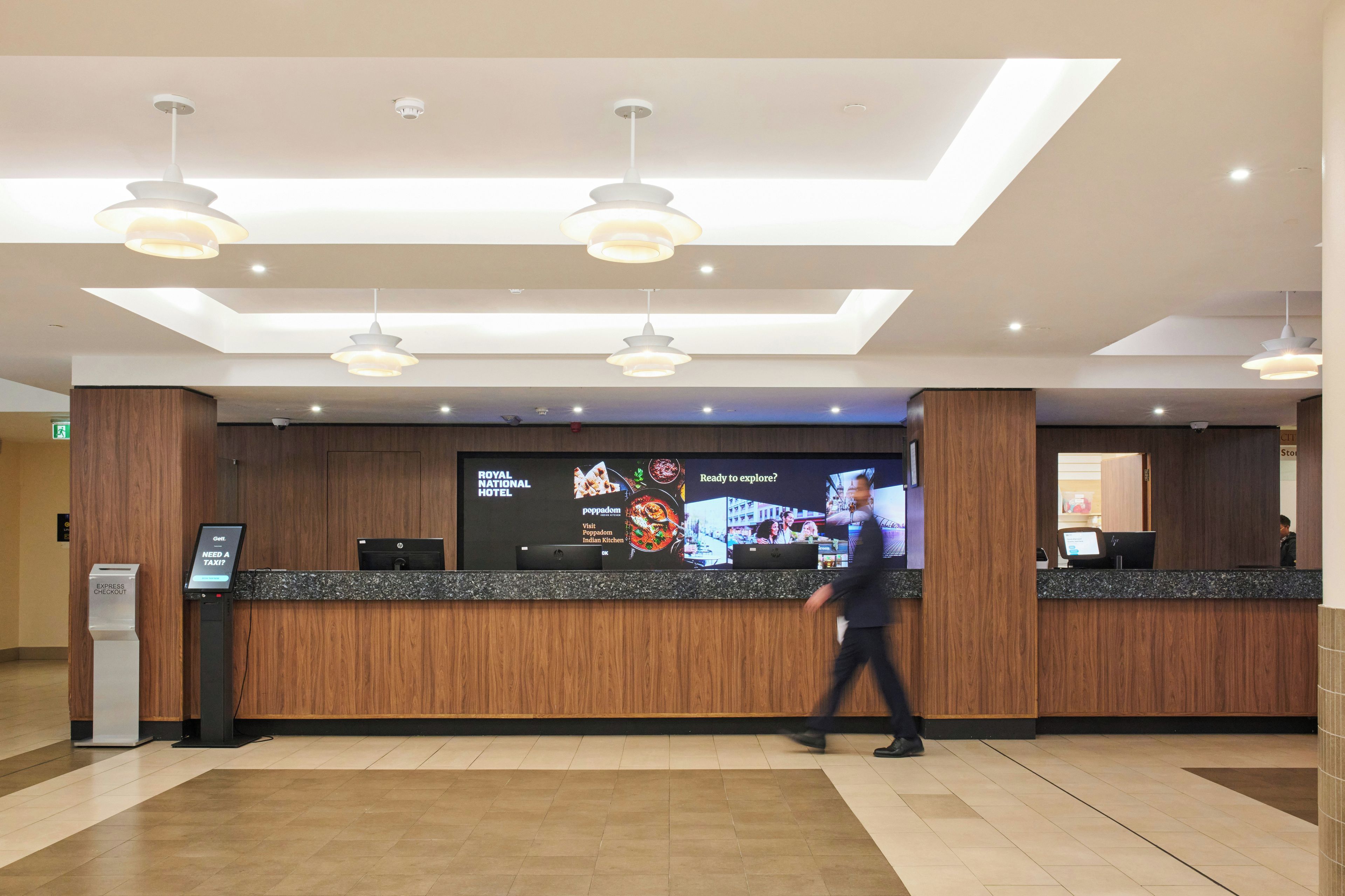 A hotel reception area with a digital display, a self-service kiosk, and a wooden reception desk.