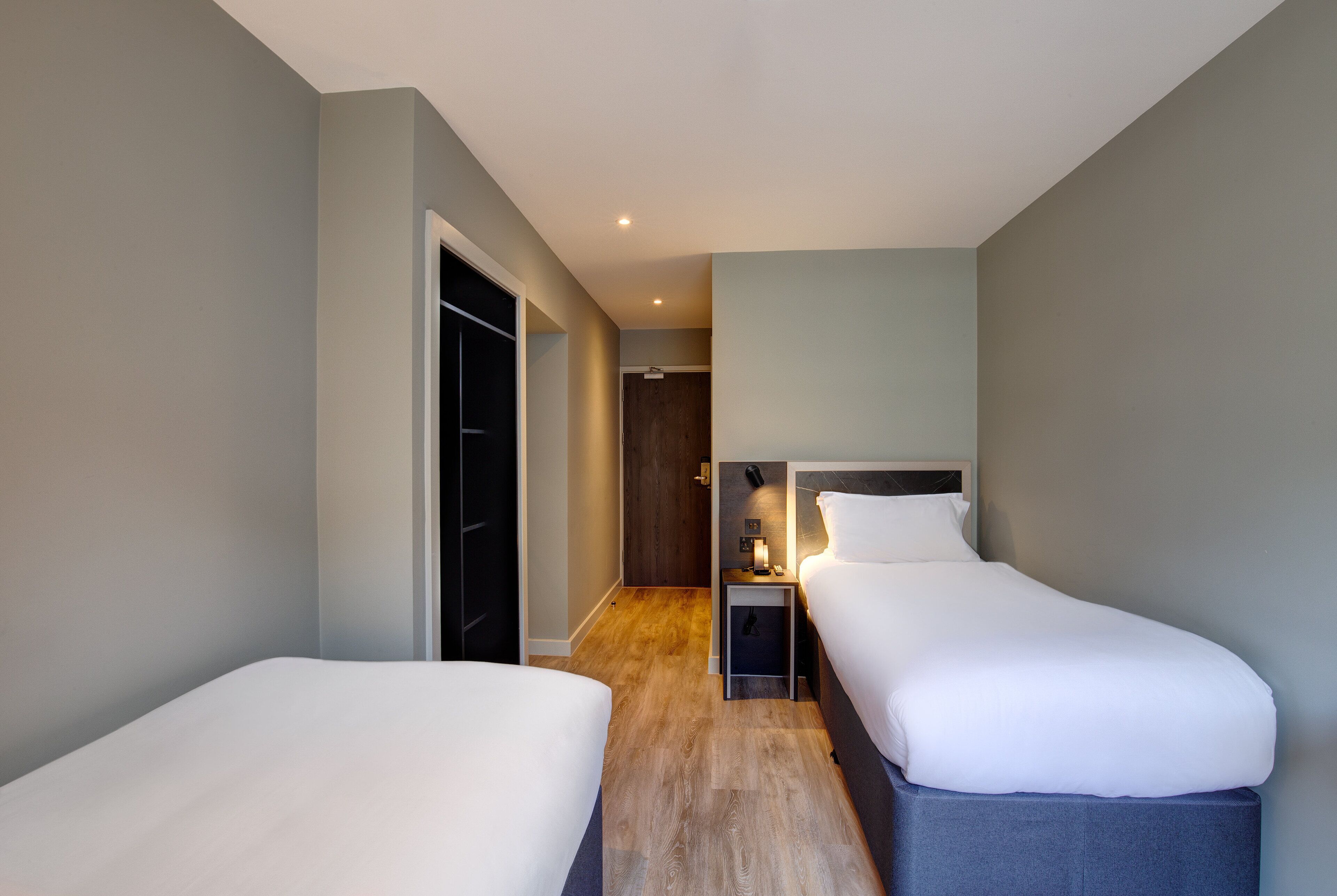 A hotel room with two single beds, a small bedside table with a lamp, a wardrobe, and a door leading to another room.