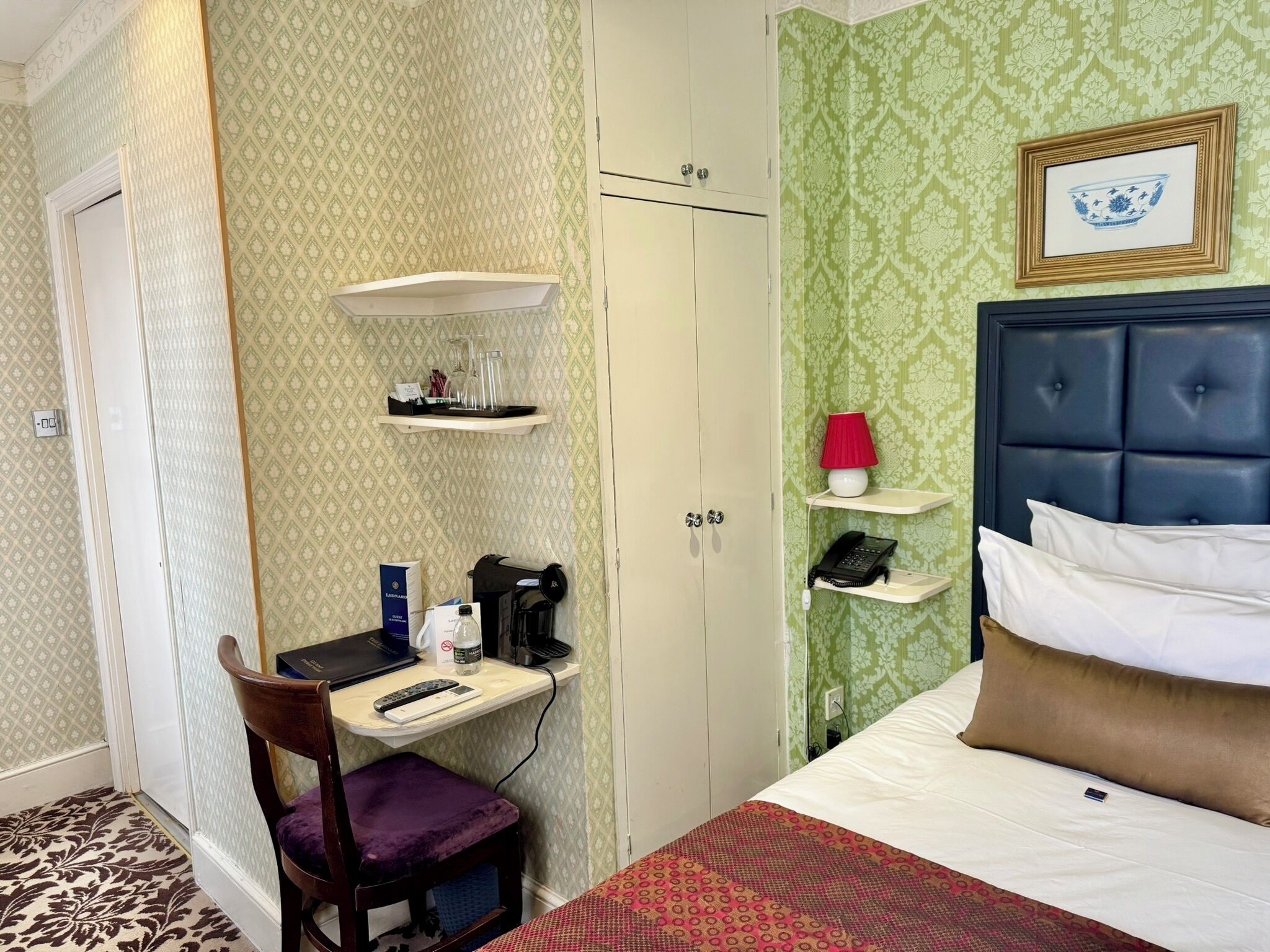 Une chambre d’hôtel comprenant un lit, un bureau avec une chaise, une armoire et une étagère avec une lampe et un téléphone.