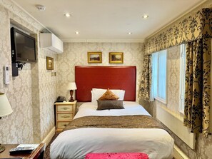 Une chambre d’hôtel avec un grand lit, un téléviseur fixé au mur, une table de chevet avec une lampe et une fenêtre agrémentée de rideaux à motifs.