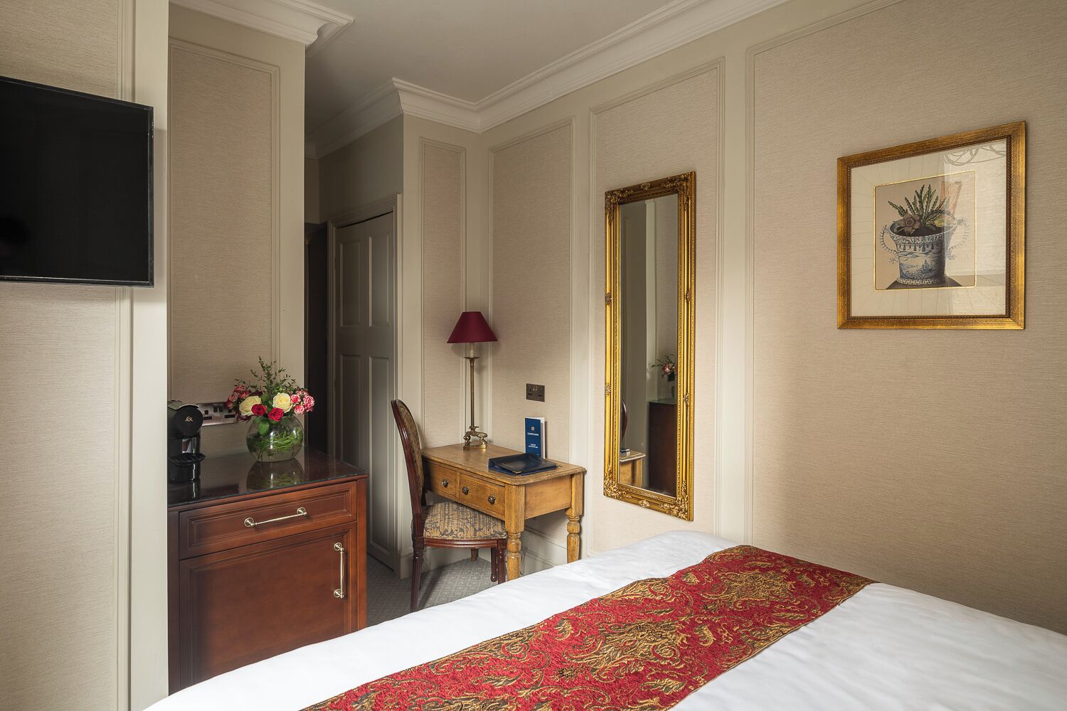 Habitación de hotel con cama, escritorio, silla, televisión, espejo y un cuadro en la pared.