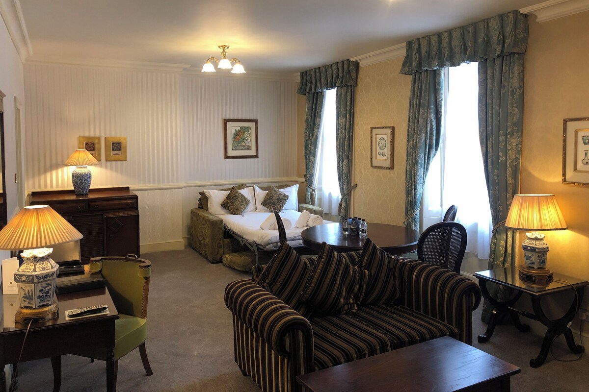 A hotel room with a bed, striped sofa, armchairs, a small table, lamps, a desk, and a window with curtains.