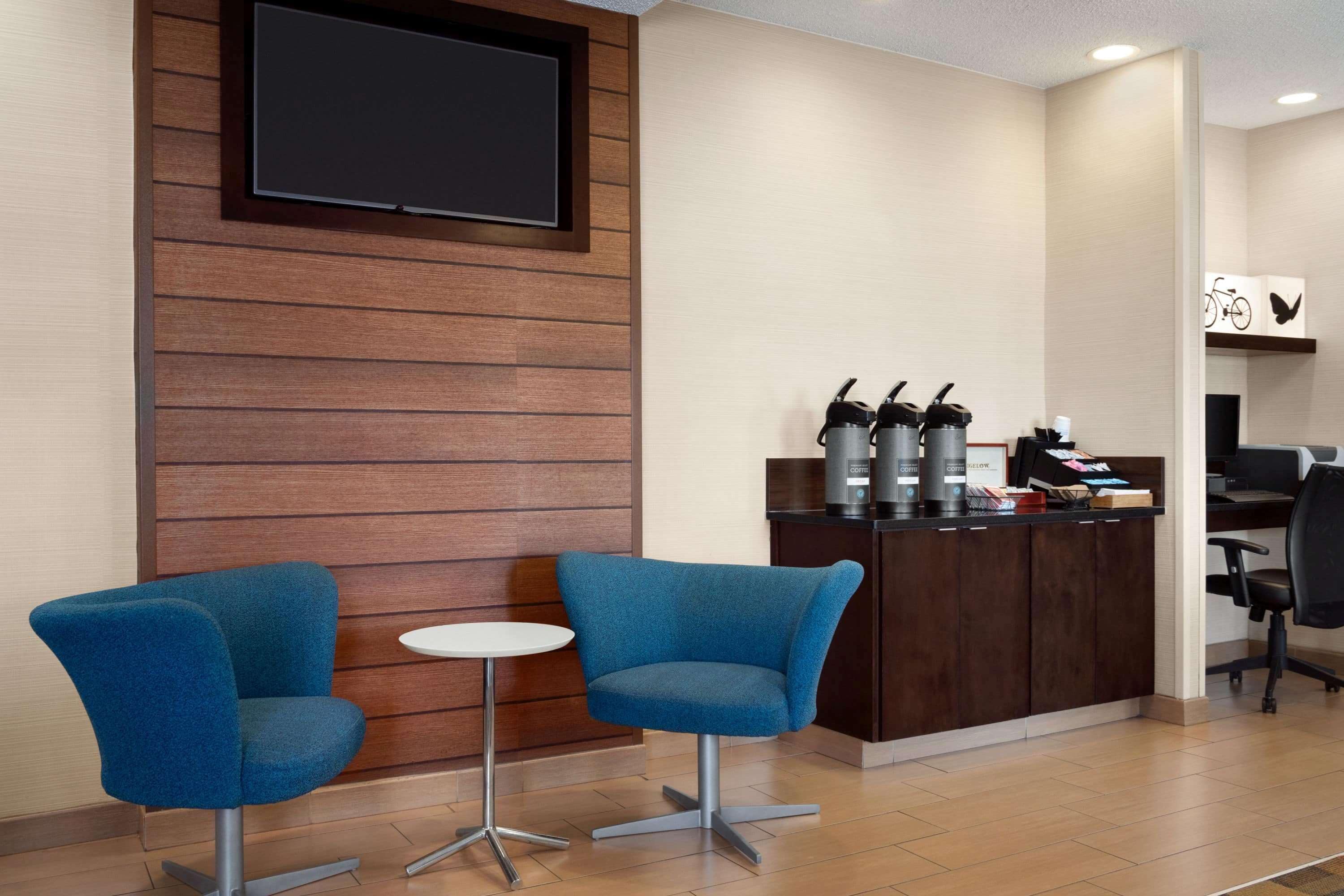 Een receptiegedeelte met twee blauwe stoelen, een kleine ronde tafel, een houten wand met een tv, een balie met koffieautomaten en een decoratie met fietsenmotieven.