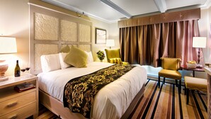 A hotel room with a large bed, a bedside table with a lamp, a chair, a small table with a bowl of fruit, and a window with curtains. -  Holland America Skagway Inn (Skagway)