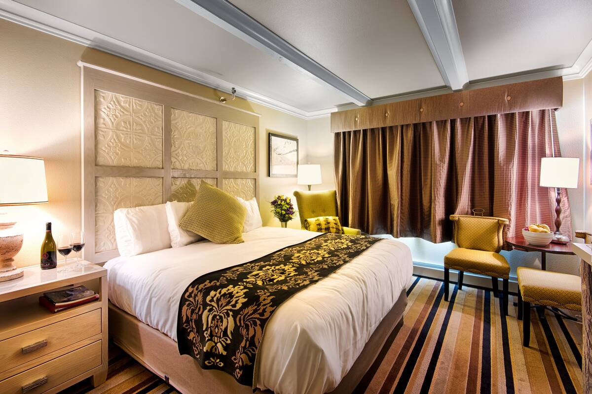 A hotel room with a large bed, a bedside table with a lamp, a chair, a small table with a bowl of fruit, and a window with curtains.