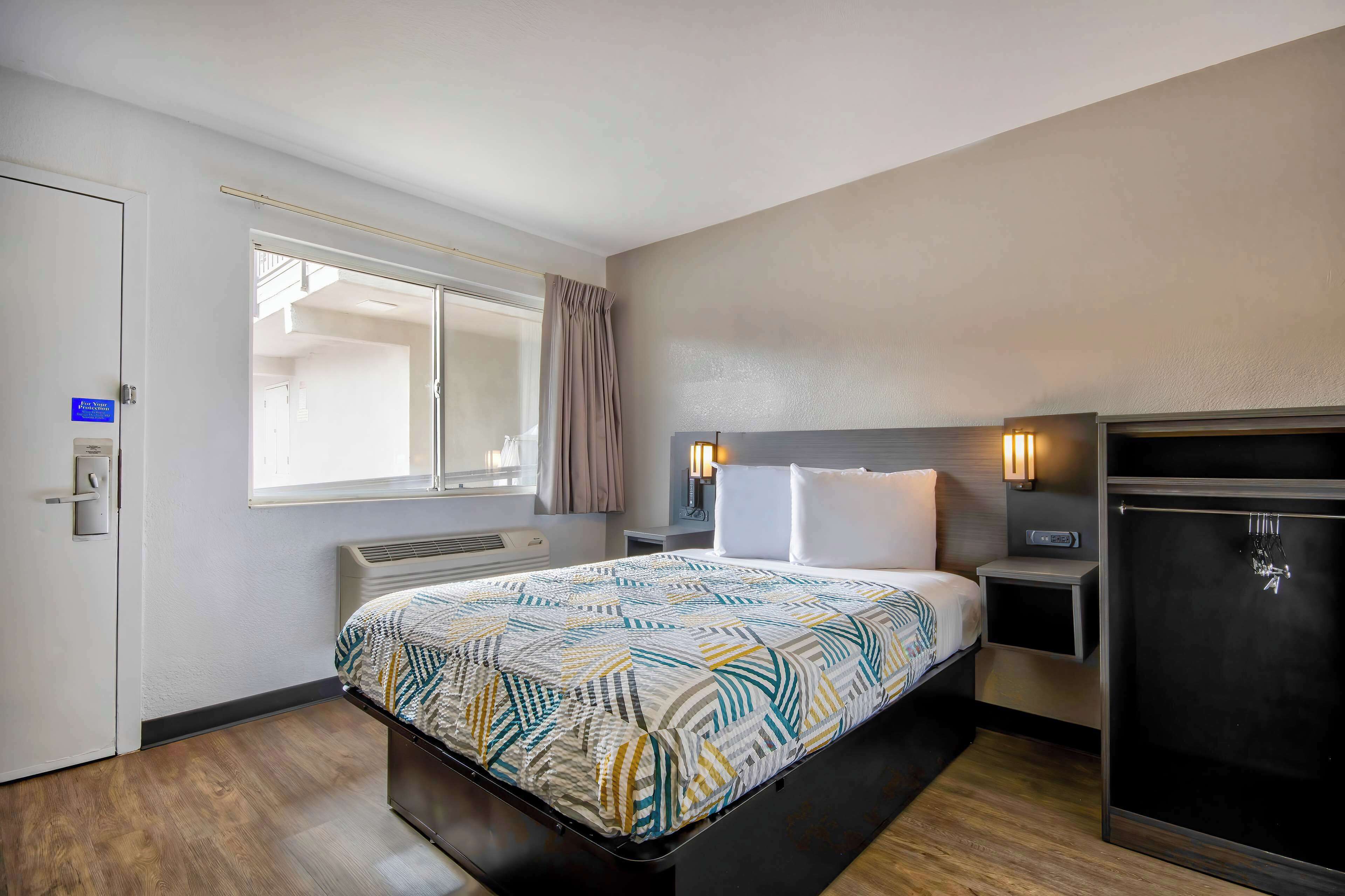 A hotel room with a bed, a bedside table, a nightstand, a wardrobe, a window with curtains, and a thermostat.