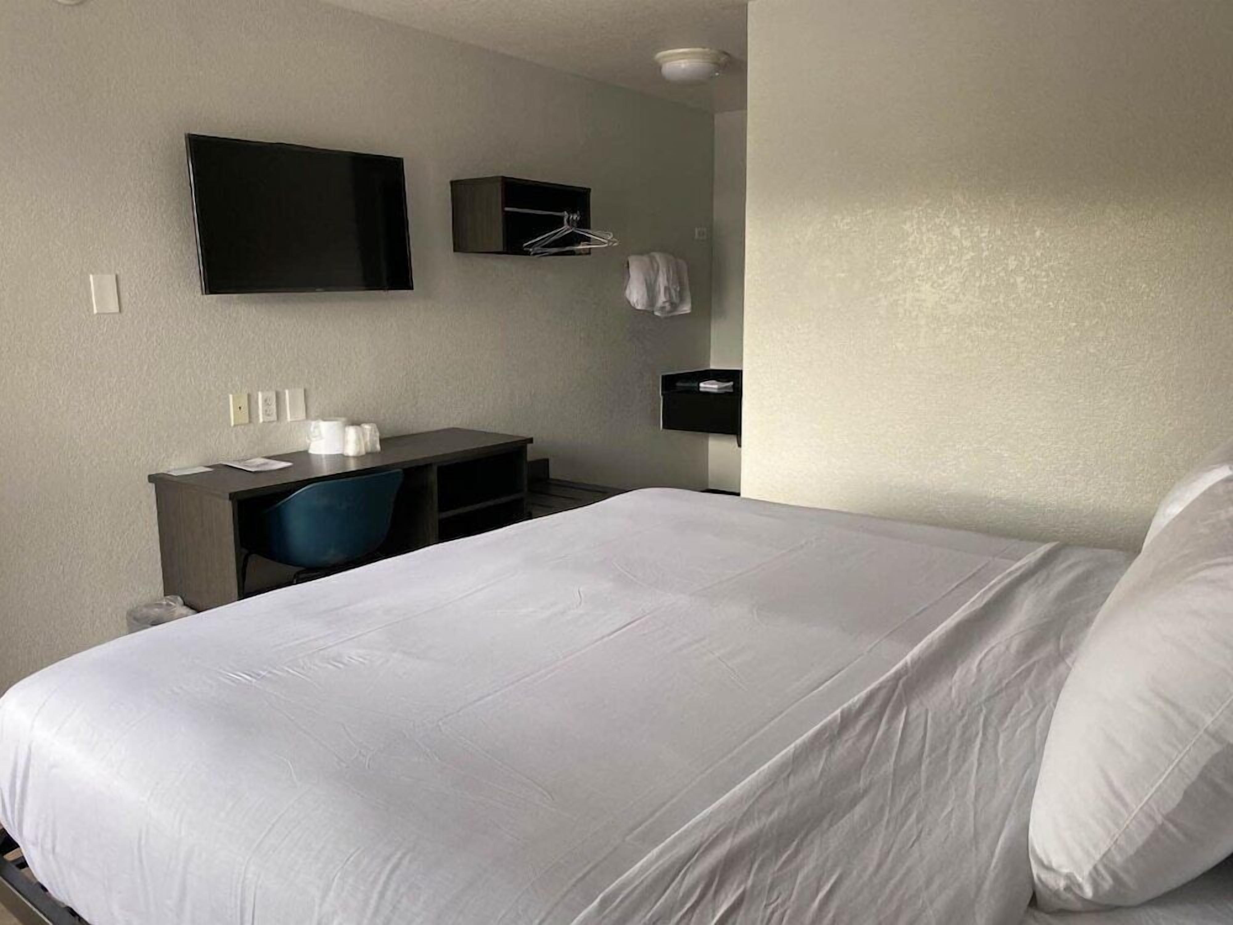 A hotel room with a bed, a desk, a chair, a TV, and a small shelf with items.