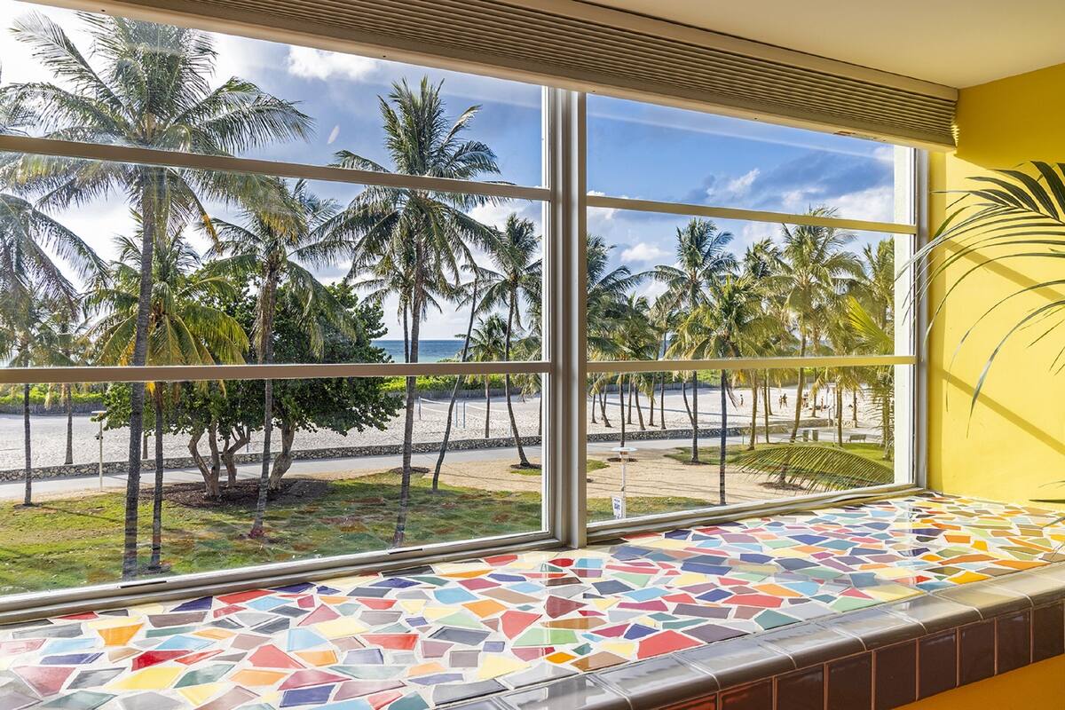 A room with a large window overlooking a beach with palm trees and a clear sky.