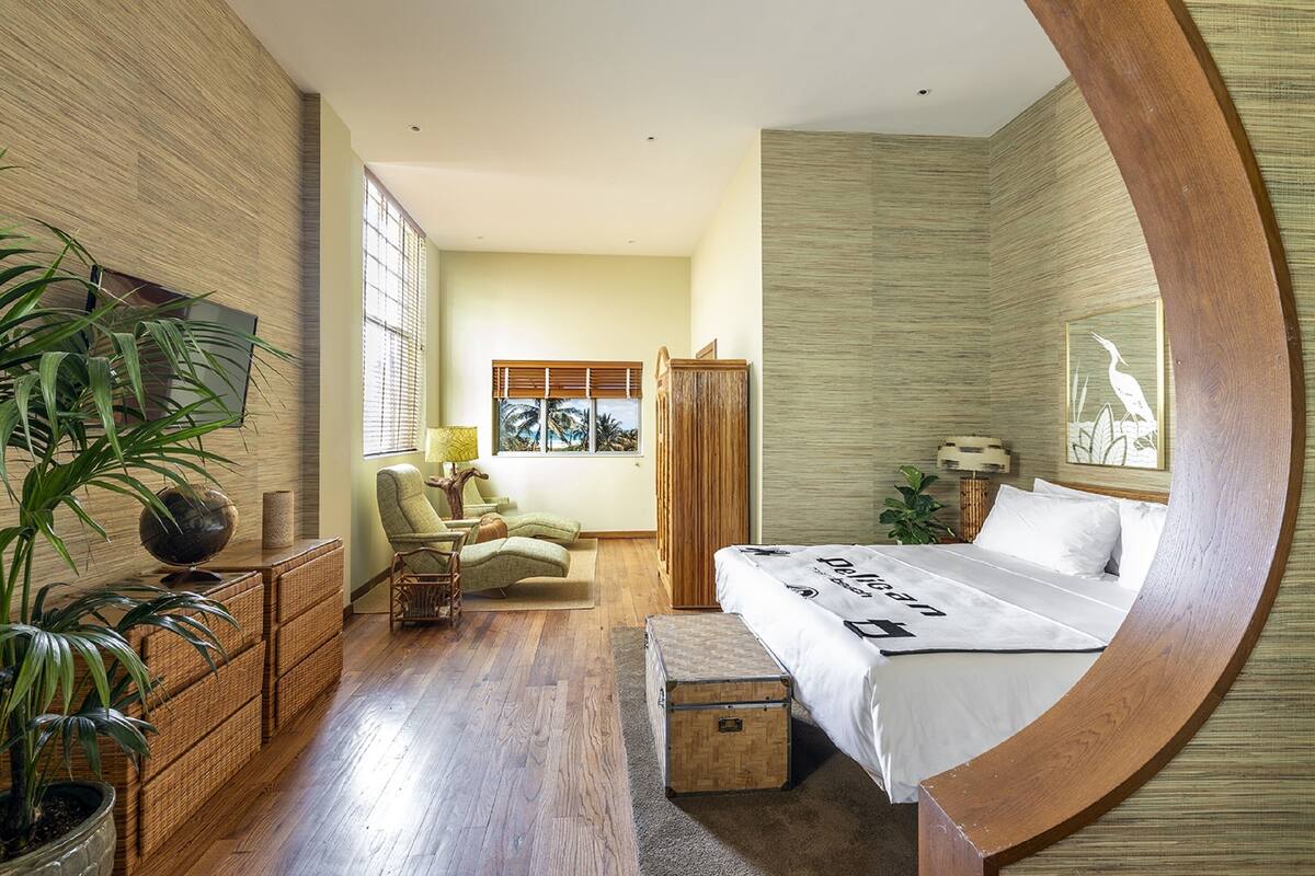 A bedroom with a large bed, a wooden wardrobe, a chair, a desk, and a large window with blinds.