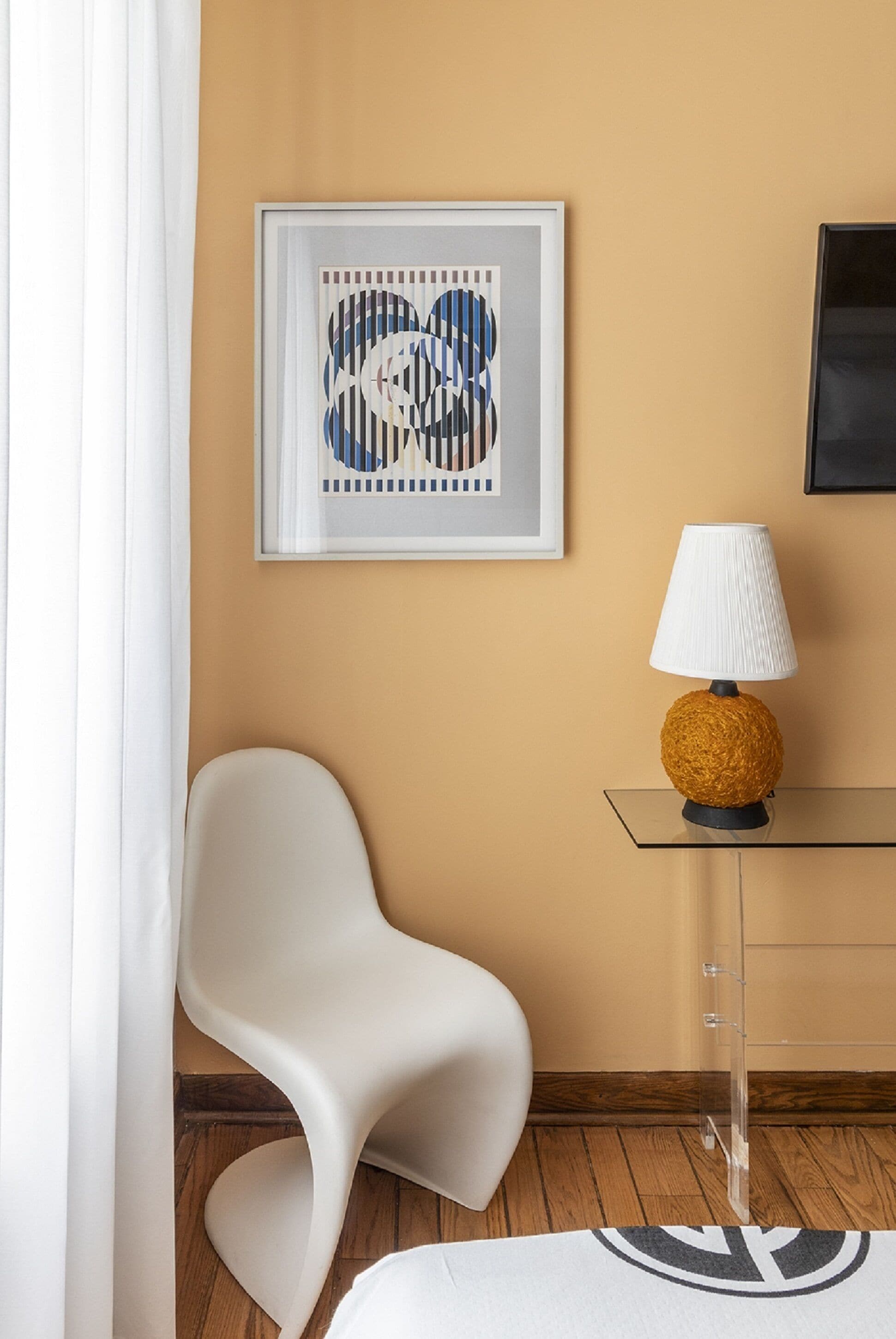 Una habitación moderna con una silla blanca, una mesa de vidrio con una lámpara y una obra de arte abstracta enmarcada en la pared.