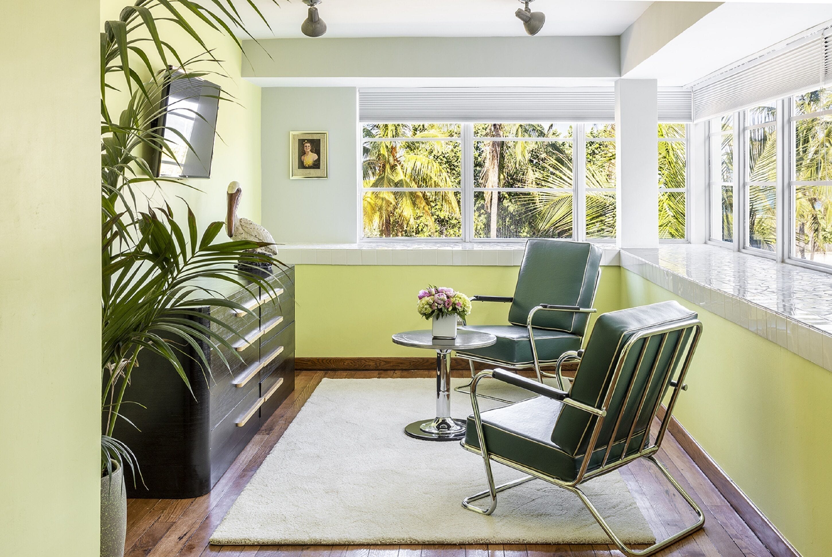 Una habitación con un ventanal grande, una silla verde, una mesita con un jarrón y una planta en maceta.
