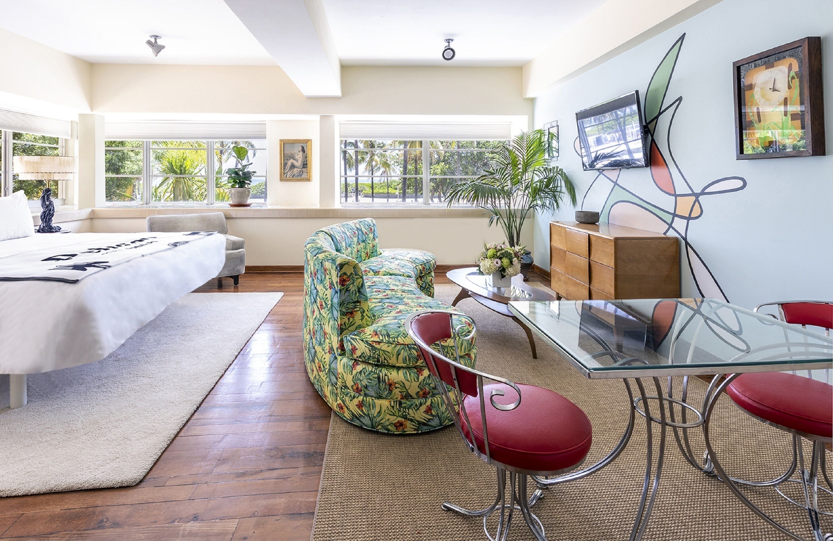 A spacious room with a bed, a sofa, a glass table, and red chairs. There are windows with a view of greenery outside.