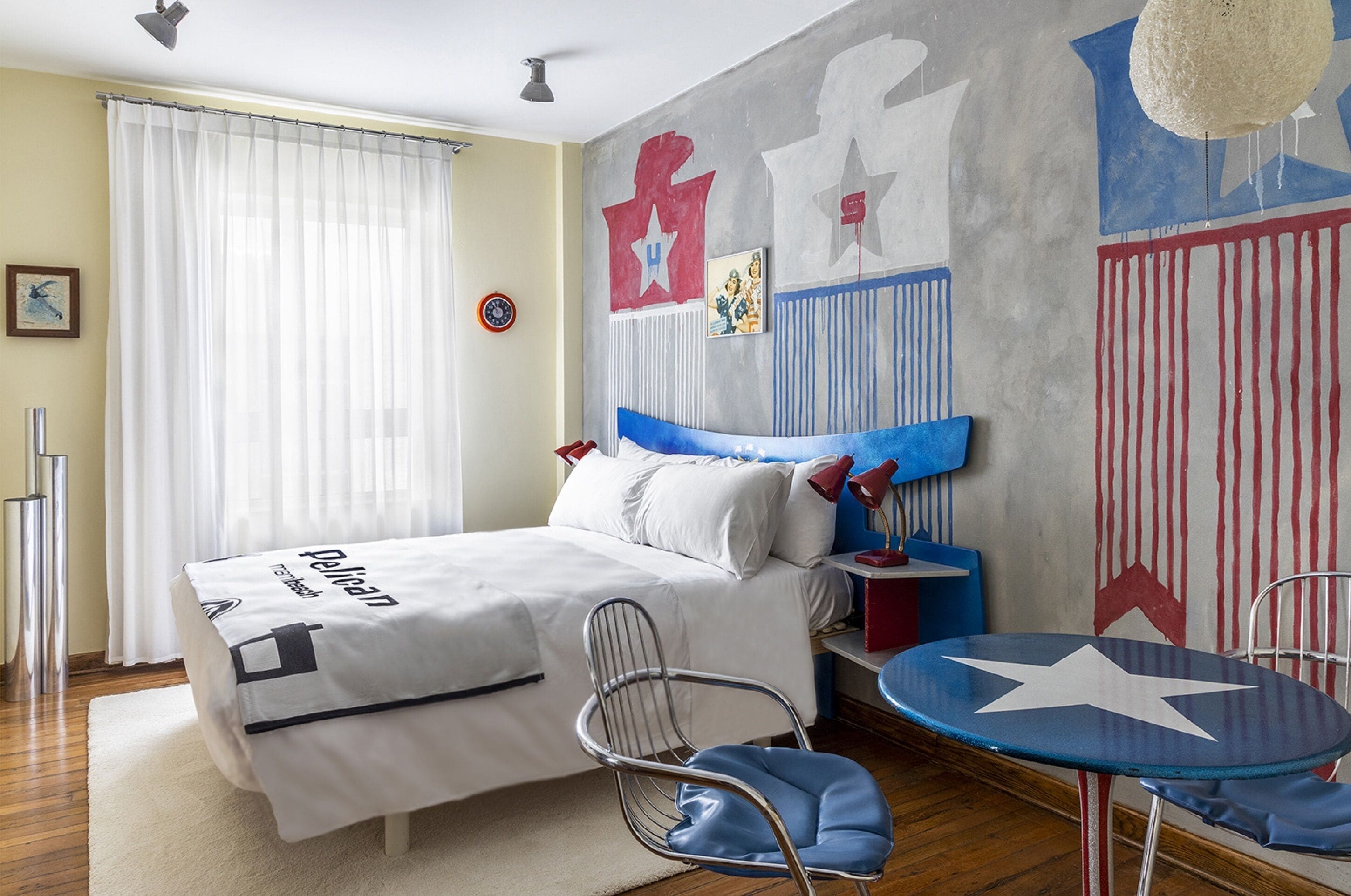 Quarto com cama, cadeira, mesinha e parede com um mural.