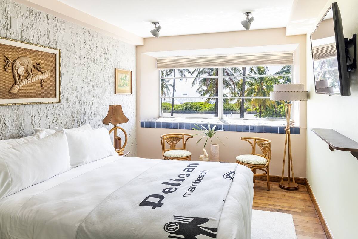 A hotel room with a bed, a window with a view of palm trees, a framed picture on the wall, and a lamp.