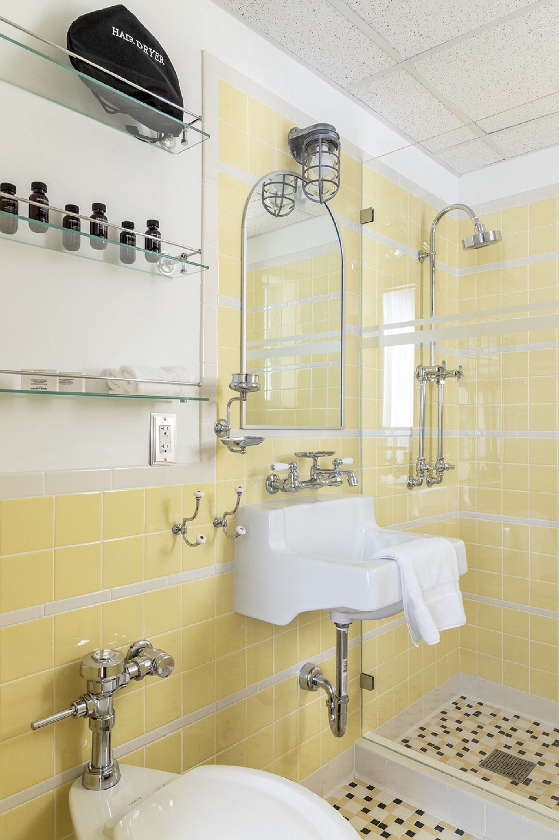 A bathroom with yellow tiled walls, a white sink, a glass shower enclosure, and a hairdryer on a shelf.