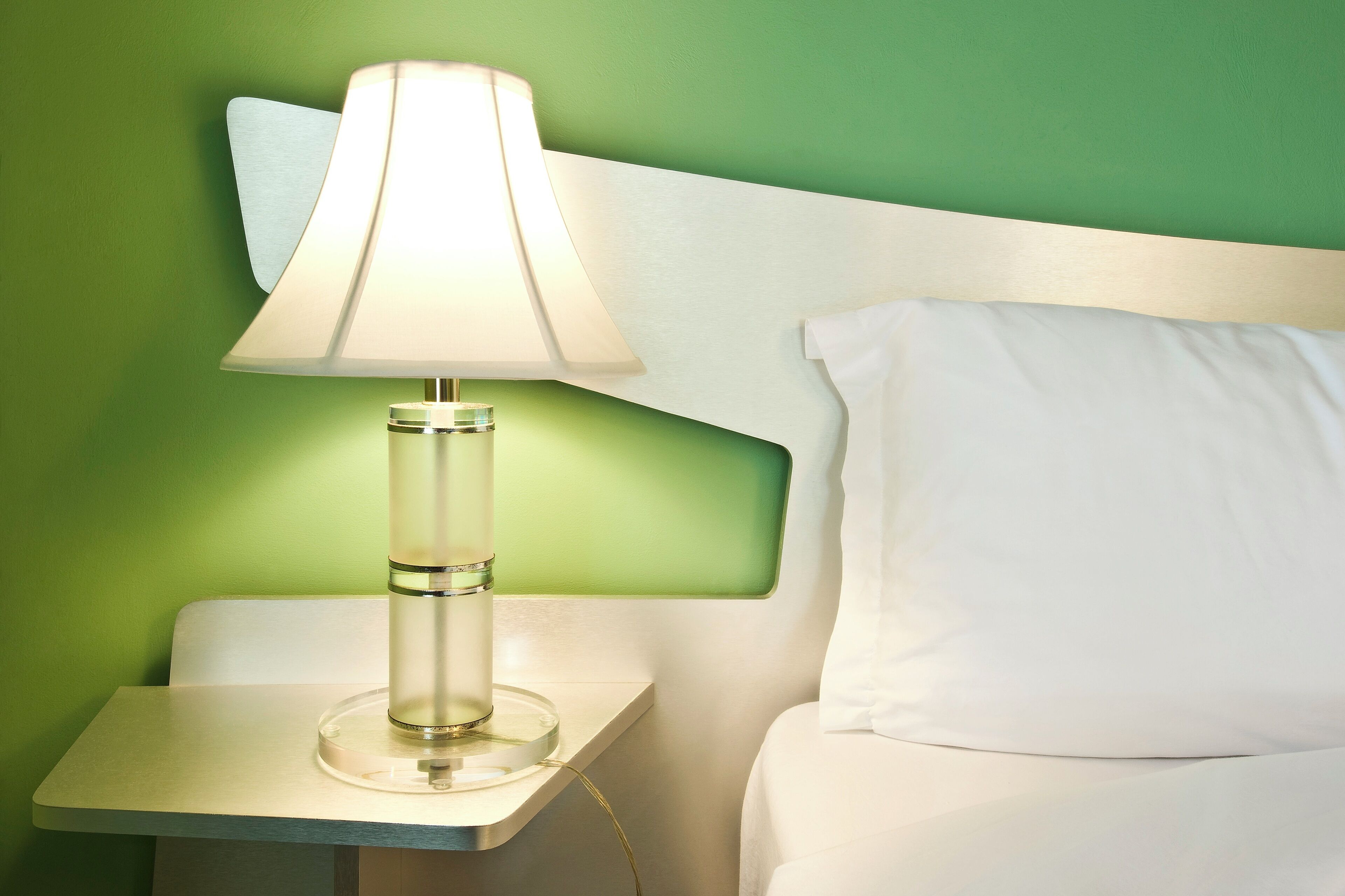 A neatly made bed with a white pillow and a bedside table with a lamp.