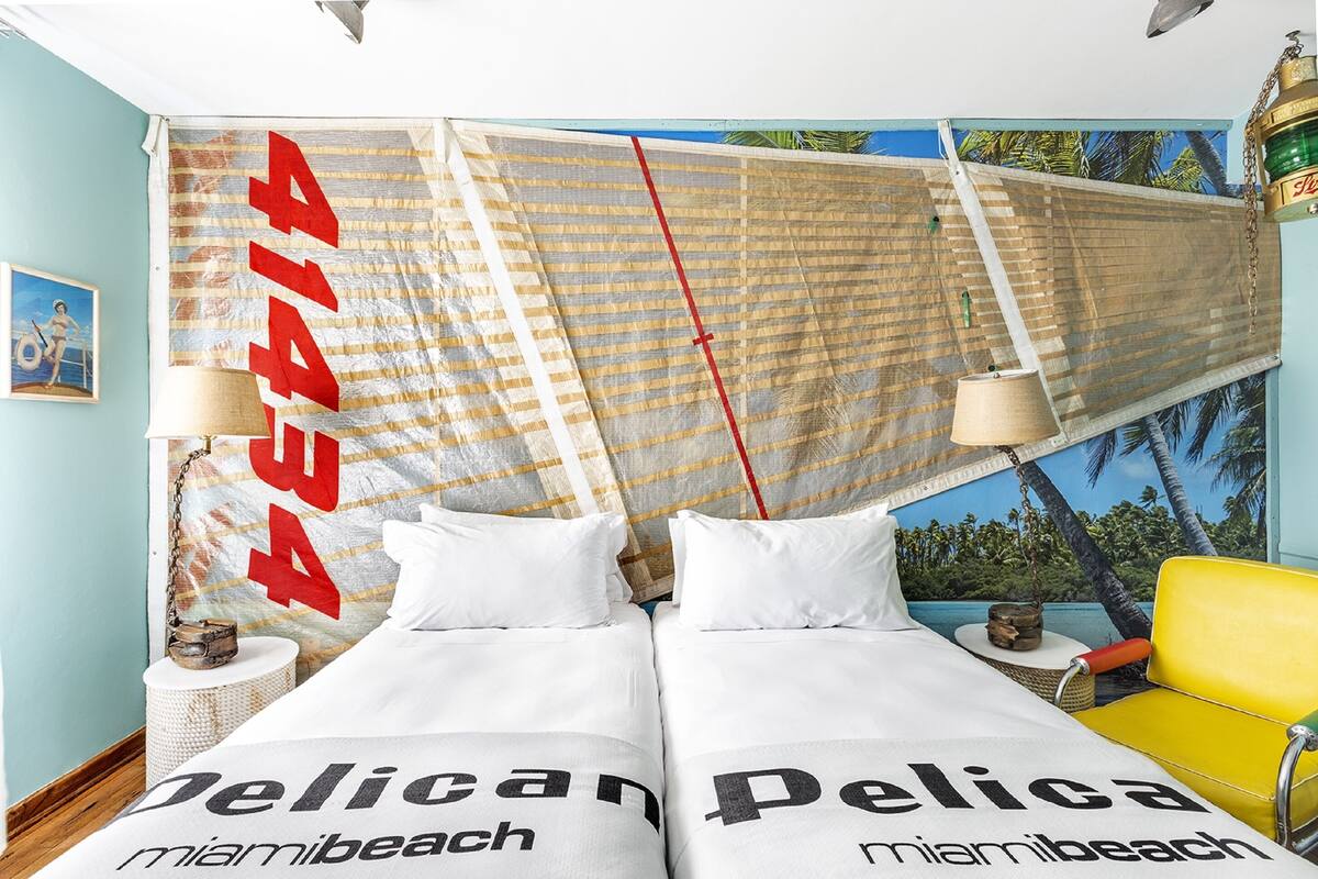 A room with two beds, each with a pillow labeled "Pelica Miami Beach." There is a yellow chair and a small table with a lamp. The room has a mural of palm trees and a beach scene on the wall.