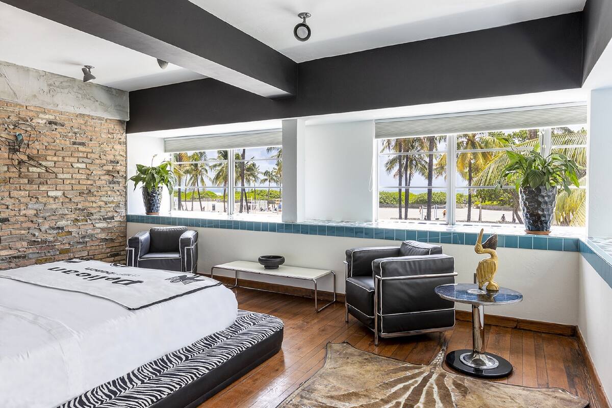 A modern hotel room with a large bed, a seating area with a table, and a view of palm trees through large windows.